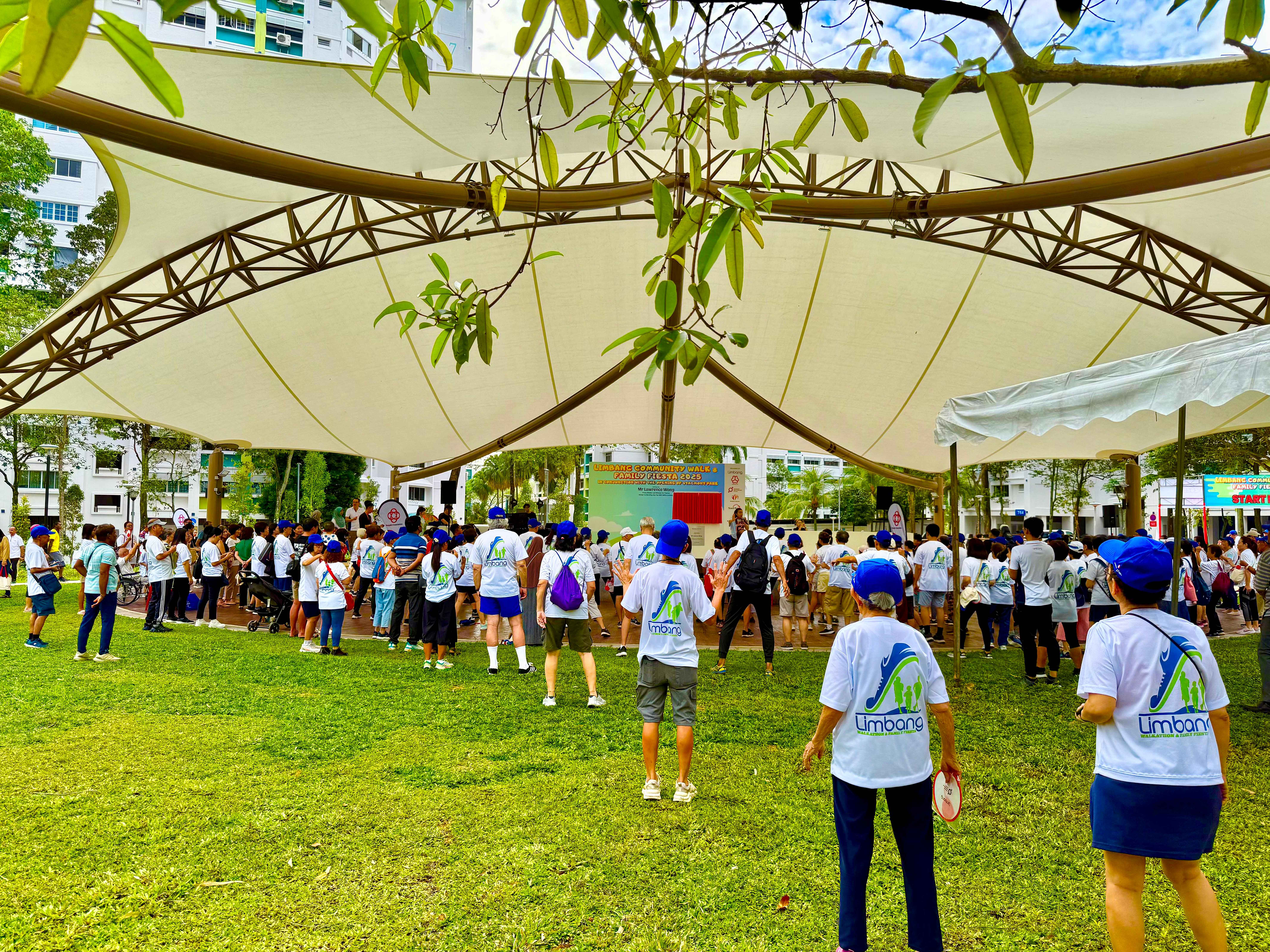 Crowd in matching white shirts gathers on grass under a large canopy, facing a stage backdrop.